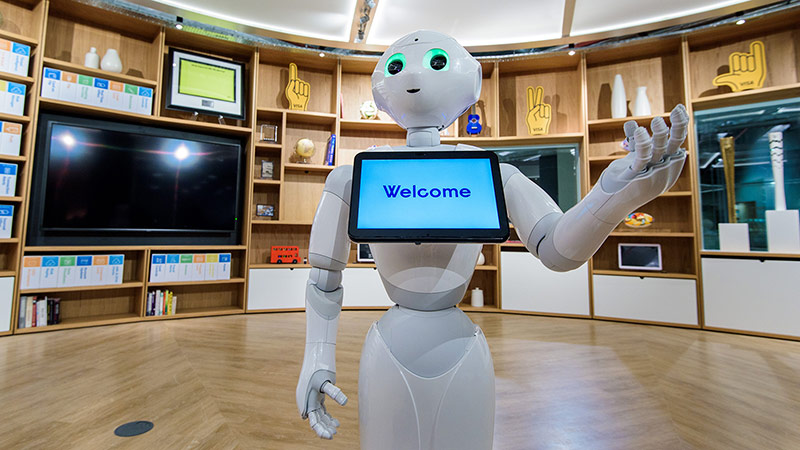 A semi-humanoid robot named Pepper standing with a tablet positioned just below its head with the message 'Welcome.'
